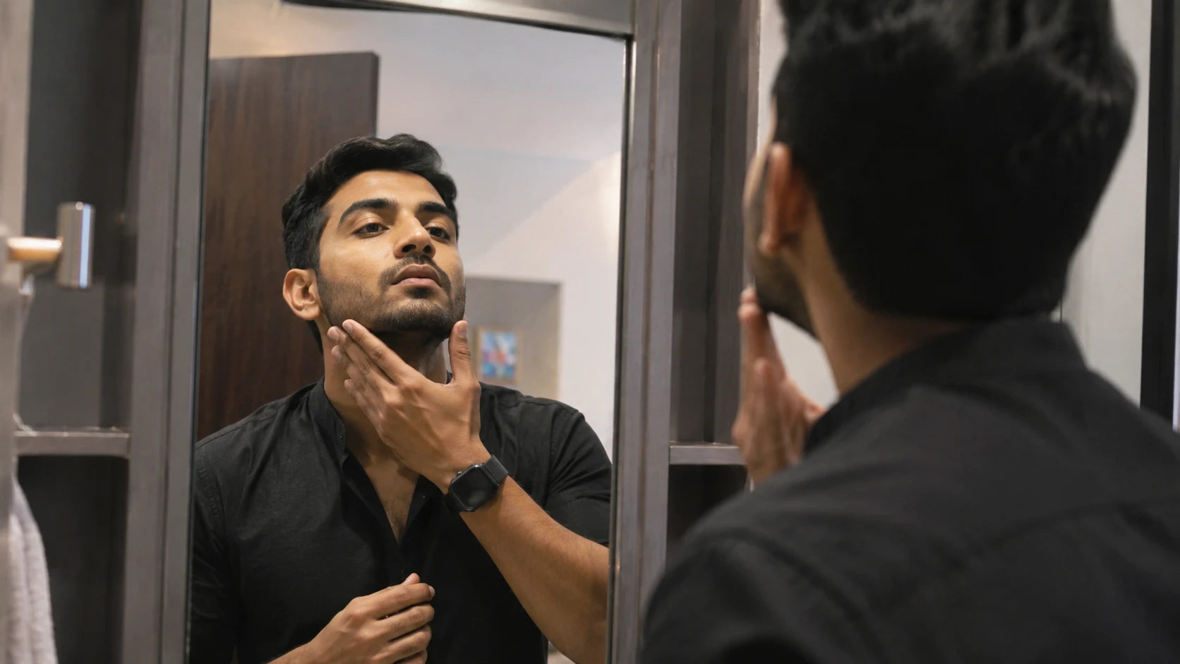 A man in a black shirt looks into a bathroom mirror while touching his chin, with his reflection and his own back visible in the frame.